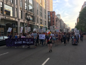 Activists march in the Boston Dyke March, 2019, holding signs that read "gay as fuck", "disability rights are an LGBTQ issue" and more