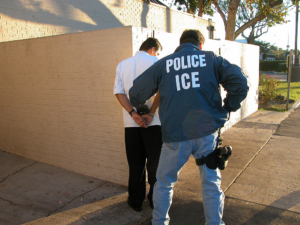 Armed ICE officer arrests man in white shirt and black pants.