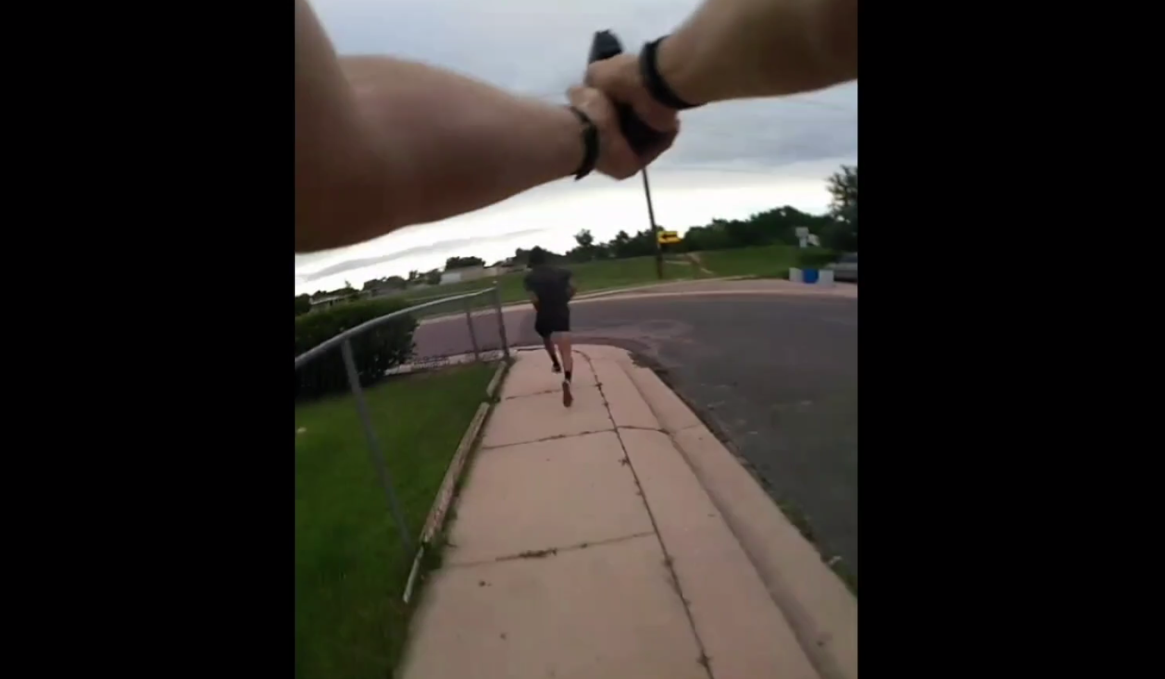 Image from badge camera of police officer shows hands holding a gun pointed at a small figure in distance running away on a path.
