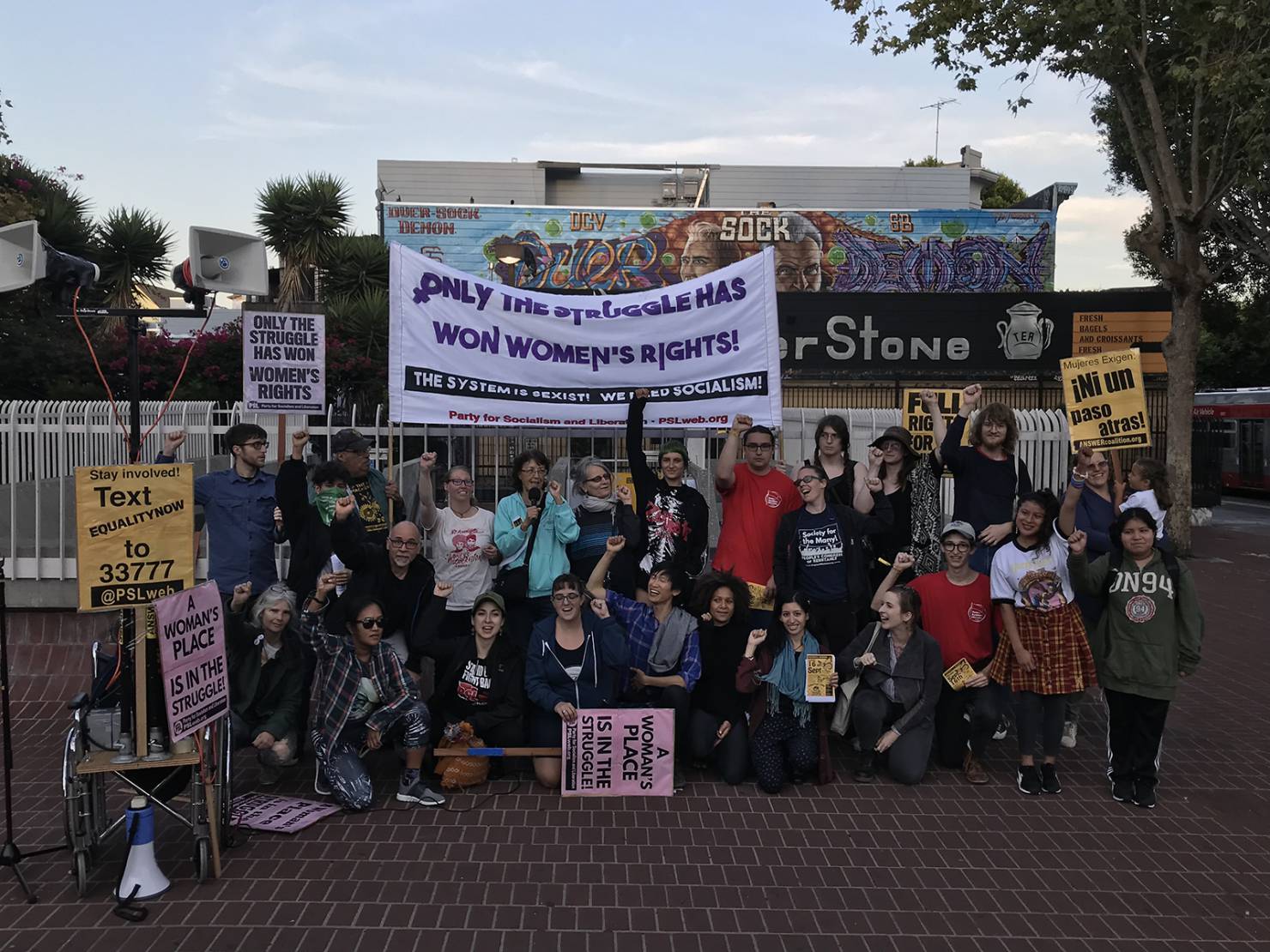 A group of activists, men and women, stand with fists raised in front of the 24th and Mission BART station in San Francisco. They hold a banner reading "Only the struggle has won women's rights!"