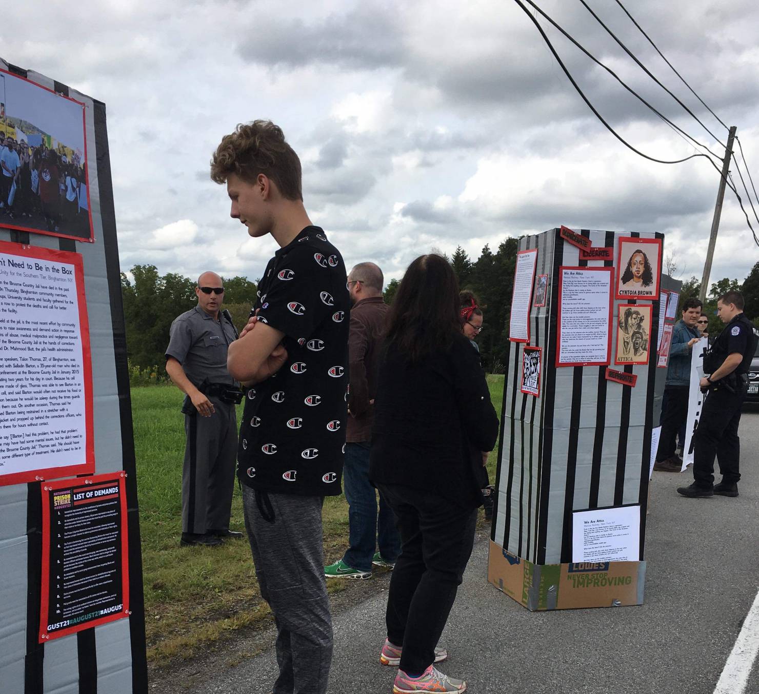 People stand on the roadside, looking at cardboard kiosks on which are placed images and typed text.