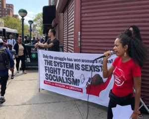A red-shirted activist holds a mic in front of a banner which reads "Only the struggle has won women's rights. The system is sexist, fight for socialism." 