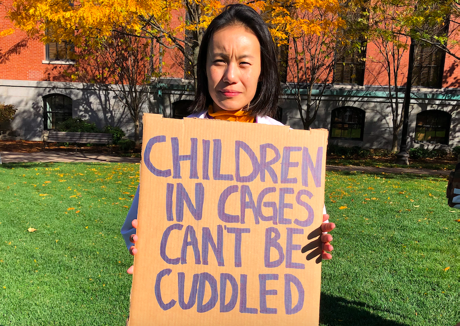 protestor wearing a lab coat holds a sign reading "children in cages can't be cuddled"