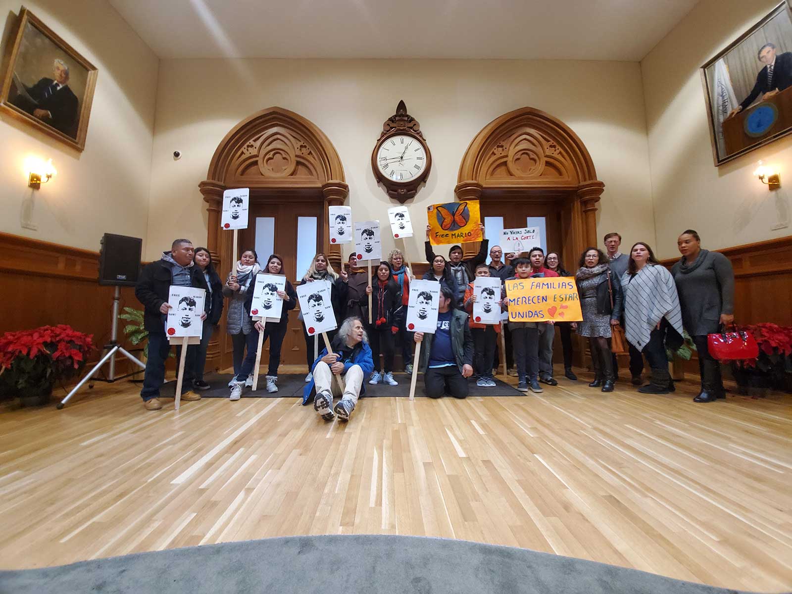 Protesters stand and sit with signs on a wooden floor.