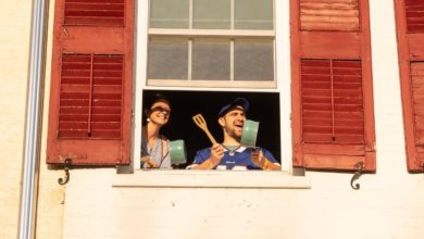 Two people stand inside a window, smiling and banging cooking pots with wooden spoons.