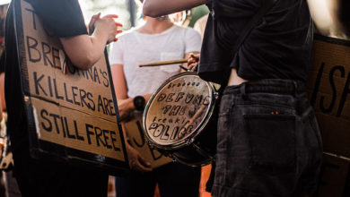 A protester holds a drum, where painted on the top it reads, "brooooo — defund the freaking police." Another protester holds a sign that reads "Breonna's killers are still free."