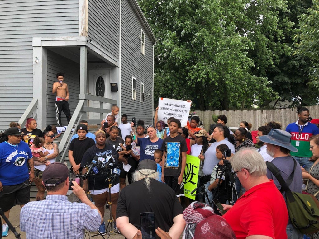 A large crowd gathers outside a grey house on Sayles Street. There are many microphones and media present. People hold signs.