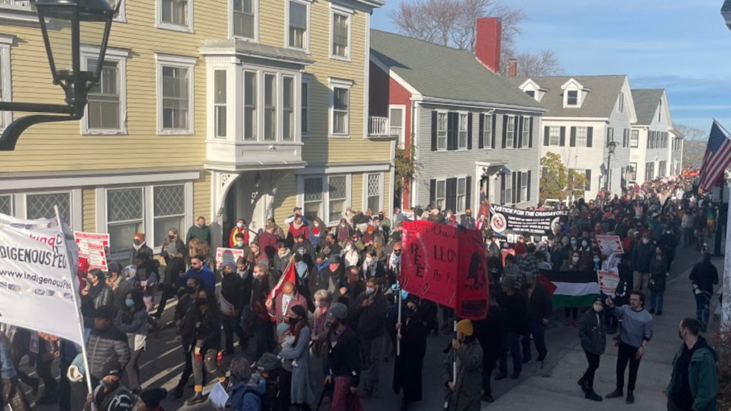 The National Day of Mourning march fills the main street in Plymouth, Massachusetts. Participants are dressed warmly and can be seen chanting. Visible at the front of the group is a banner that reads "#sinkingcolumbus: Indigenous Peoples Day Now!" Another banner reads "In the Spirit of Crazy Horse, Free Leonard Peltier Now!" Beside that is a Palestinian flag, all held by participants.