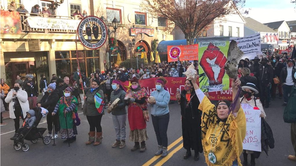 As the march passes restaurants and gift shops on colonized Wampanoag land (Plymouth), all banners are visible at the front of the crowd. Another banner carried by a participant features a symbol of a red fist. In front of this banner is Chali'naru Dones, a Taino-Boriqua woman in traditional Taino clothing. She holds aloft a conch shell and a bird's wing attached to a leather handle, her expression stern and proud. Individuals around her also wear clothing traditional to their Indigenous cultures.