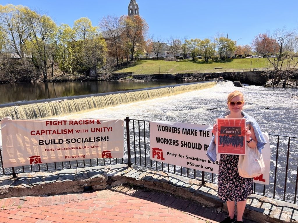 A participant takes a photo in front of PSL banners with the Slater Mill waterfall as a backdrop on May Day 2022, at Slater Mill, Pawtucket, Rhode Island.